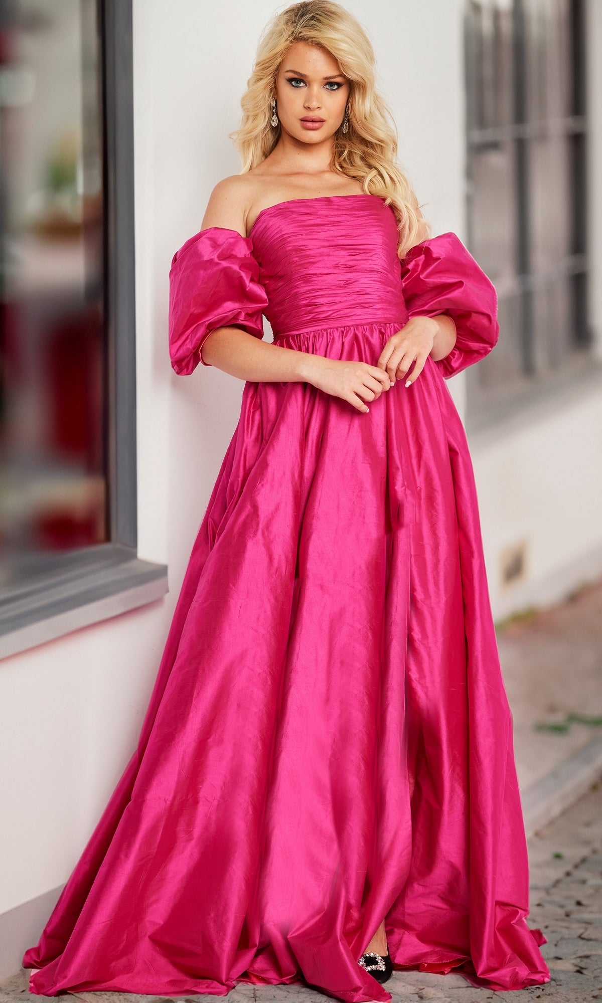 A woman stands outdoors in a bright pink Jovani Puff-Sleeve Long Prom Ball Gown 24099, leaning slightly against a wall.